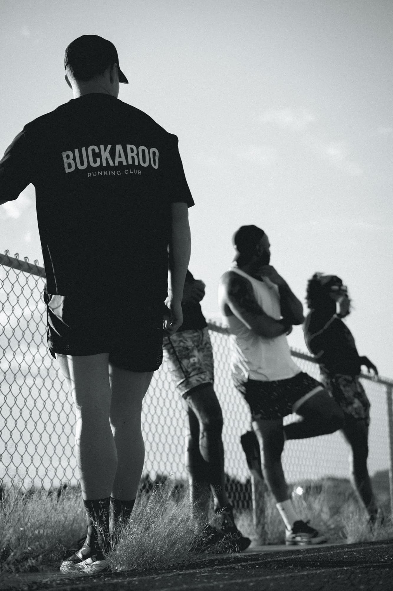 Runners in Buckaroo Running Club gear stretching against a fence on a track at sunset, in black and white
