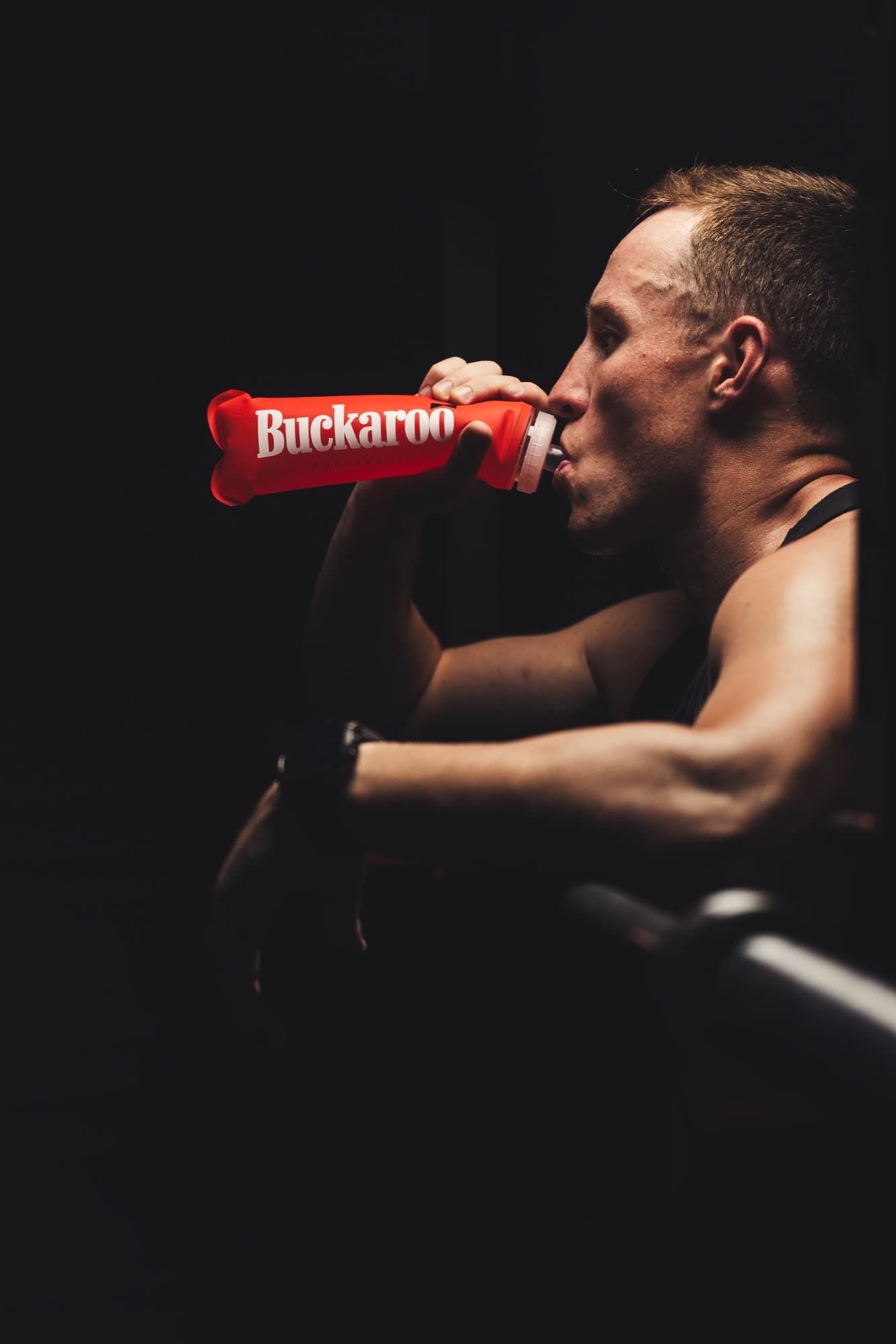 An athlete in a dark gym drinking from a red Buckaroo bottle between efforts